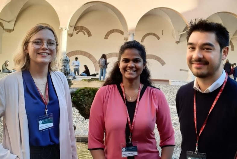 Three engineers [Jess, Hasini, Michele] stand in a courtyard. They are smartly dressed and have conference lanyards around their necks.