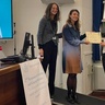 Three formally dressed women stand in front of a presentation screen. Jess, a blonde woman in a white shirt and dark trousers, is being presented with a certificate. All subjects are smiling towards the camera.