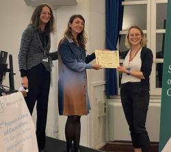 Three formally dressed women stand in front of a presentation screen. Jess, a blonde woman in a white shirt and dark trousers, is being presented with a certificate. All subjects are smiling towards the camera.