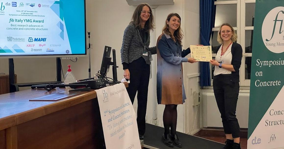 Jess recieved award for best research advances in concrete and concrete structures Three formally dressed women stand in front of a presentation screen. Jess, a blonde woman in a white shirt and dark trousers, is being presented with a certificate. All subjects are smiling towards the camera.